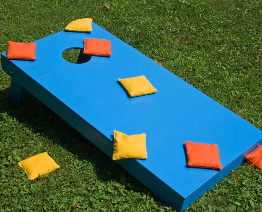 Bright blue cornhole board on grass with yellow and orange bean bags scattered on and around it.